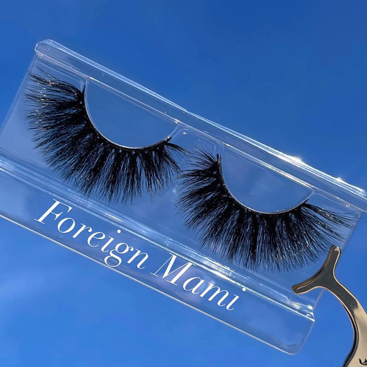A set of mink false eyelashes displayed on a clear plastic case with the brand name 'FOREIGN MAMI' written below, against a blue sky background.
